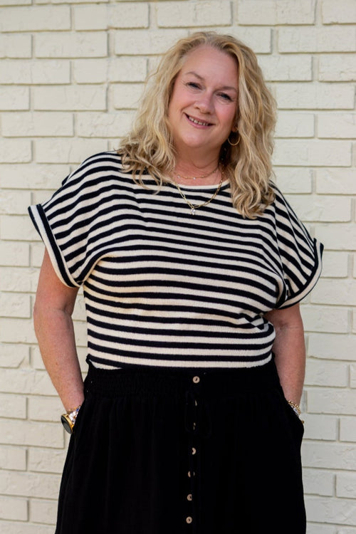 Woman wearing a black and white striped top and black skirt standing against a white brick wall.
